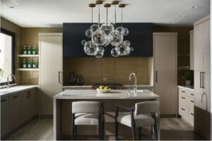 Modern luxury kitchen featuring a central island with a sleek waterfall marble countertop, paired with two plush upholstered stools. A sculptural glass bubble chandelier with brass accents hangs above, serving as the room's dramatic focal point. Matte taupe cabinetry surrounds the space, including integrated appliances and open shelving stocked with glass bottles. A minimalist chrome faucet rises from the island, while the backsplash showcases warm, glossy taupe tiles under soft natural light. The overall palette is refined and earthy, with a balance of modern geometry and inviting textures.